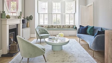 A bright living room with hardwood floors, white walls, and large windows with open white shutters. In the center of the room is a grey crushed velvet rug with a mint green and white circular coffee table and two matching mint green crushed velvet chairs. To the right is a blue velvet couch with blue and green cushions, and to the left is a white mantelpiece with a log burner, wicker baskets, and assorted vases.