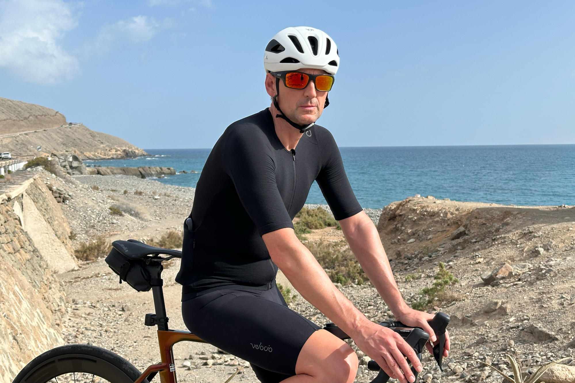 man sitting on the top tube of an orange bike wearing black shorts, white helmet and black jersey on a gravel beach