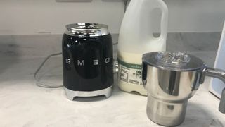 smeg milk frother beside the stainless steel jug with a carton of milk behind it