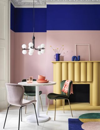 pink dining room with blue painted picture rail, yellow fire place, white round dining table, globe light and pink and green chairs