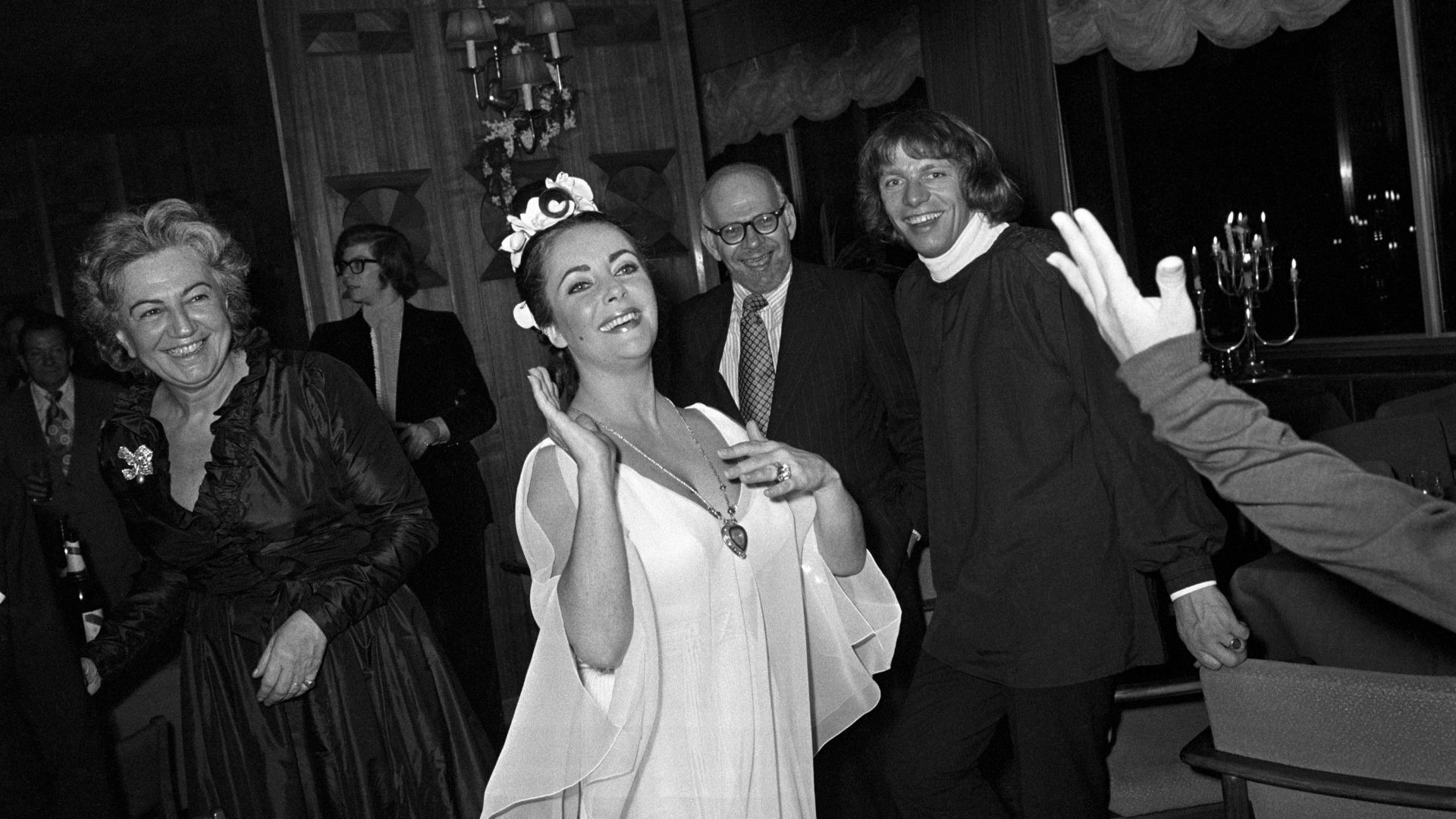 Elizabeth Taylor hitting a balloon up in the air during her post-birthday dinner party. She wears a white crepe chiffon Grecian dress designed by Edith Head with seven white orchids in her hair designed by Alexandre of Paris. Around her neck she wears a present from her husband Richard Burton the "Taj Mahal", a heart-shaped, lemon diamond pendant inscribed with Indian love poetry suspended on a gold and ruby chain by Cartier.