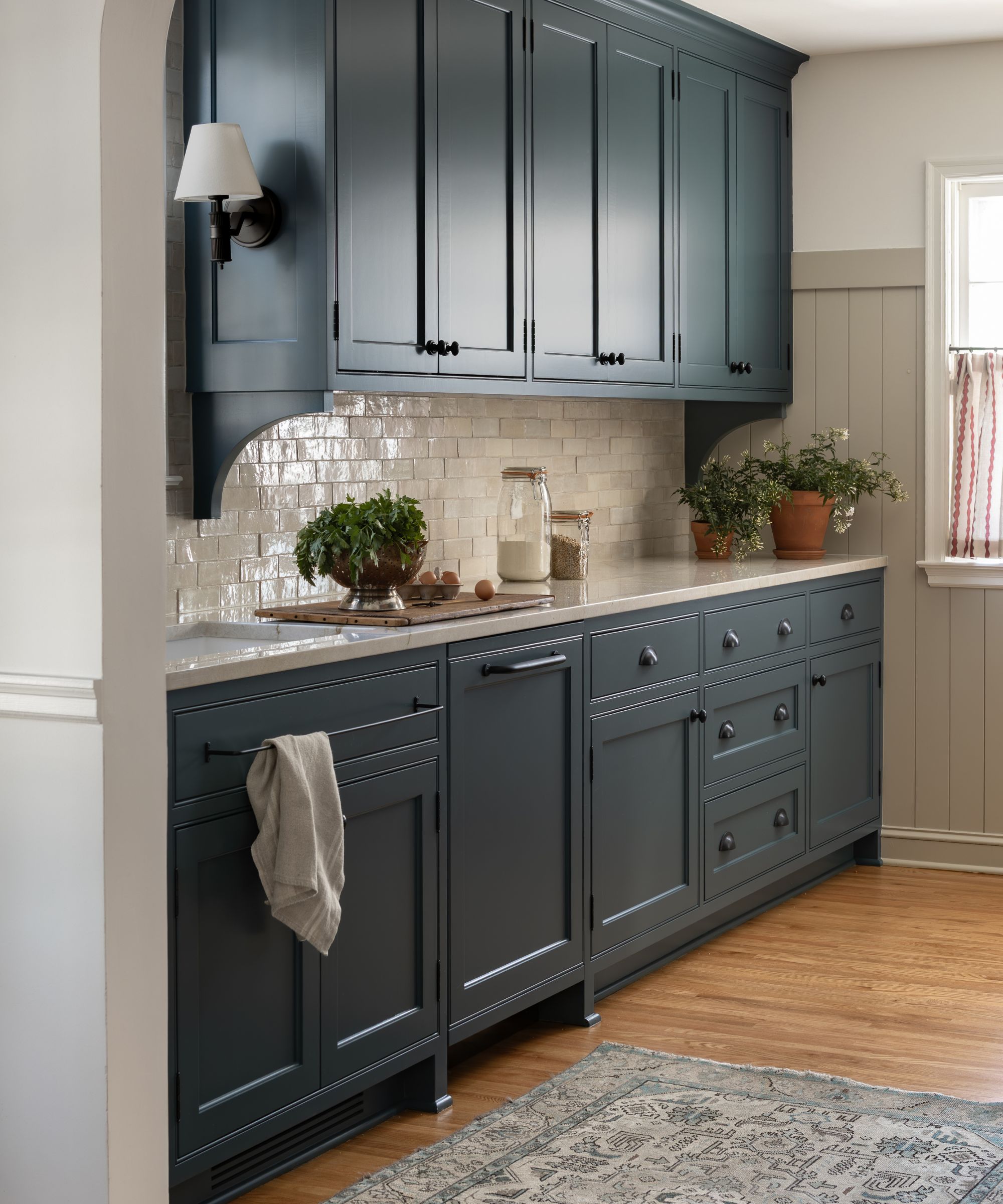 small galley kitchen painted navy blue with white zellige subway tiles, a vintage style grey rug, wall sconces and a striped cafe curtain