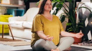 A woman sits cross legged on a yoga mat with her hands resting upwards on her knees. Behind her we see a tall, leafy plant, a sofa and a coffee table.