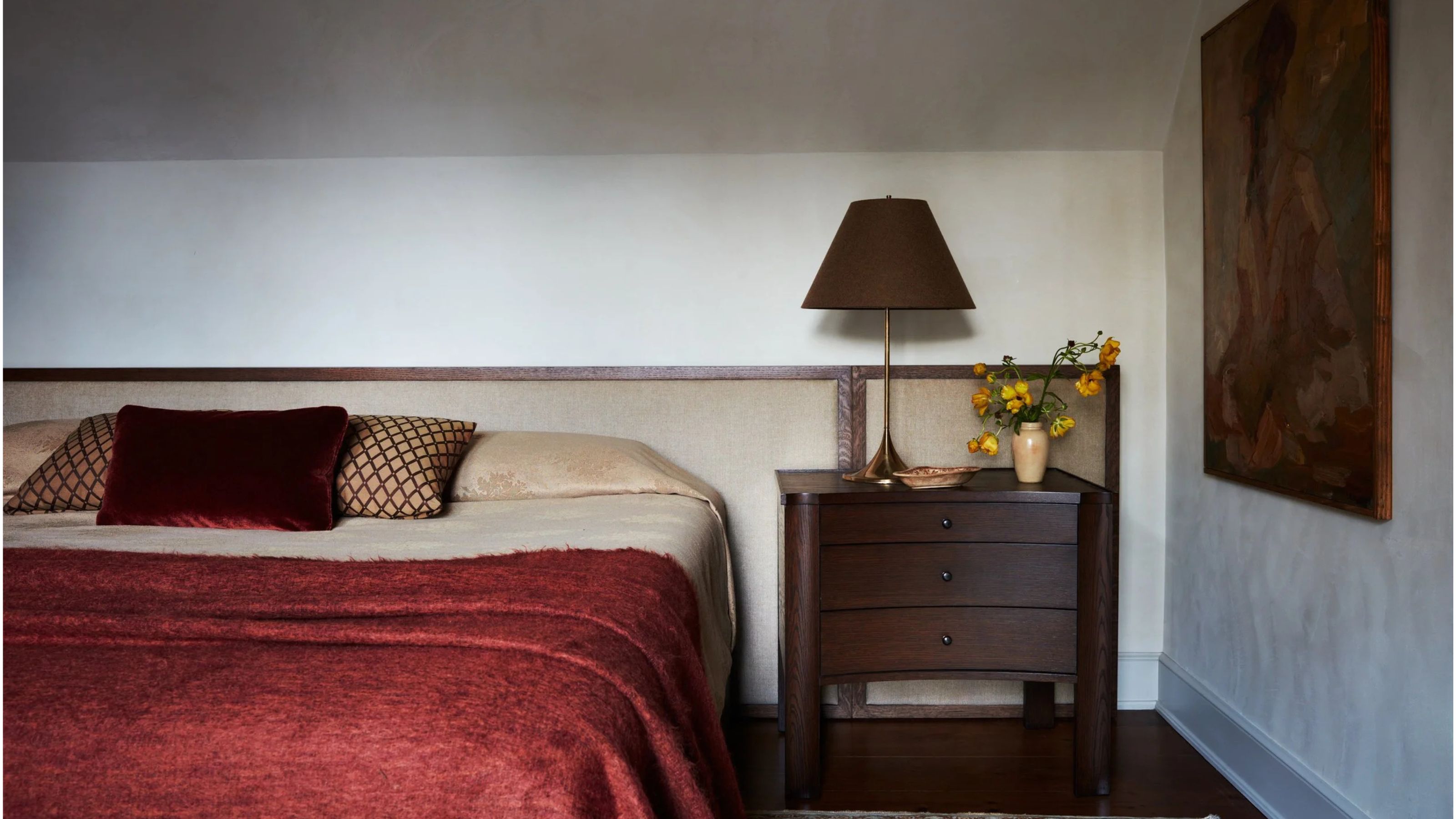 image of a traditional bedroom with angled white ceilings and white walls. There is a beige bed with an upholstered extended headboard and a wooden side table next to it. The bed has beige and red bedding.