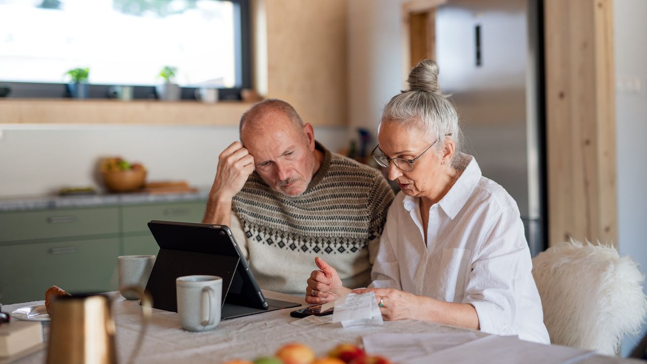 A retired couple look tense as they look over bills at their kitchen table.