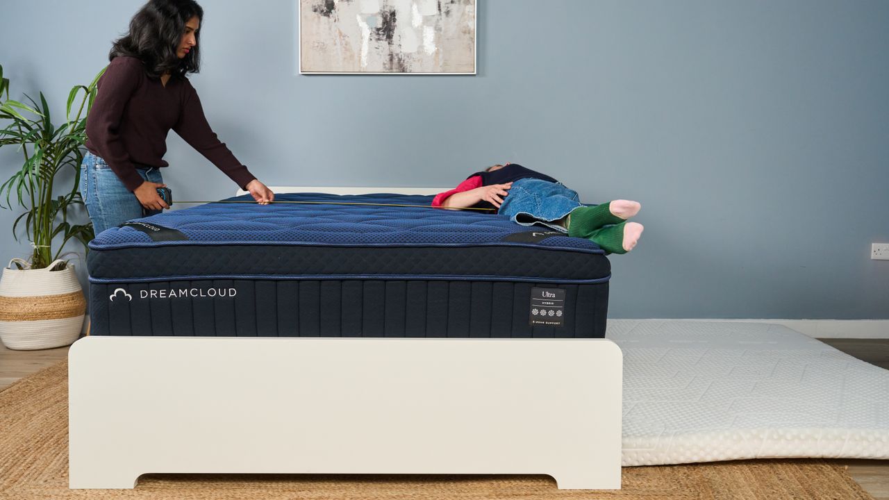 The image shows dark haired mattress tester standing next to a dark blue mattress with a measuring tape. On the opposite side of the bed, a second mattress tester slowly rolls off the side. This roll off test determines how sturdy the bed's edge is.