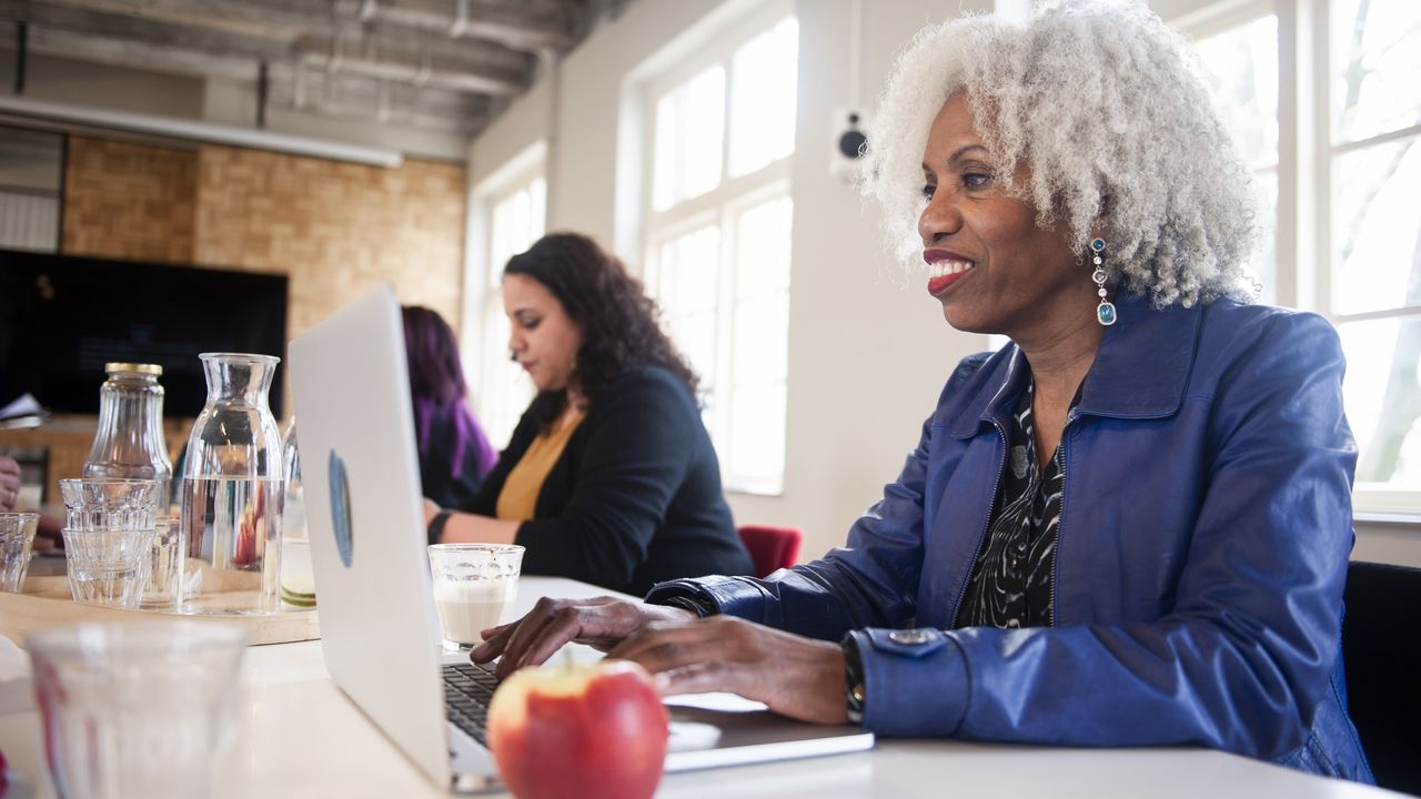 A smiling older woman works in an office with others.