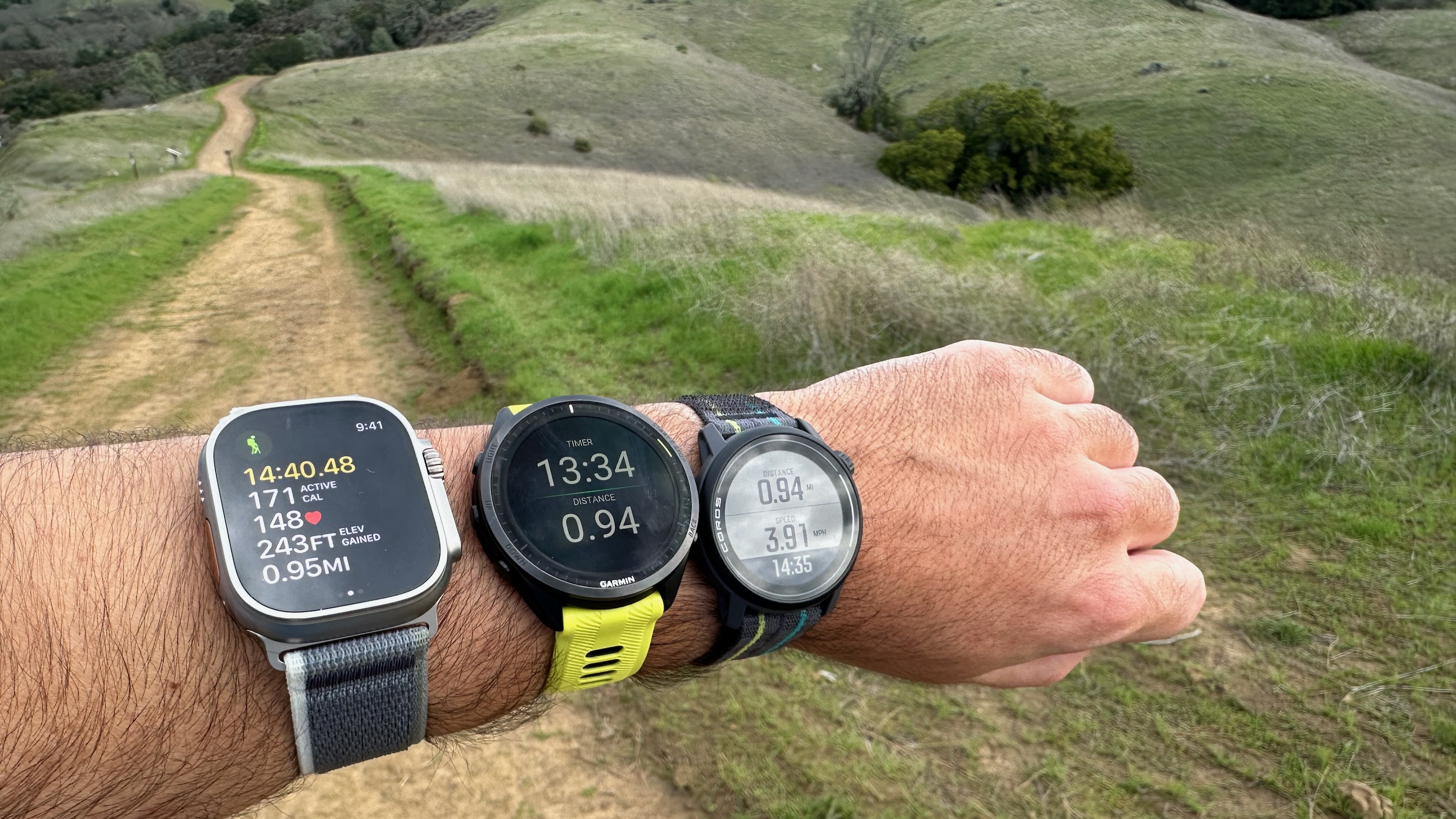 The Apple Watch Ultra 2, Garmin Forerunner 965, and COROS PACE 3 all worn on one wrist at the base of the Mount Diablo Summit Trail.