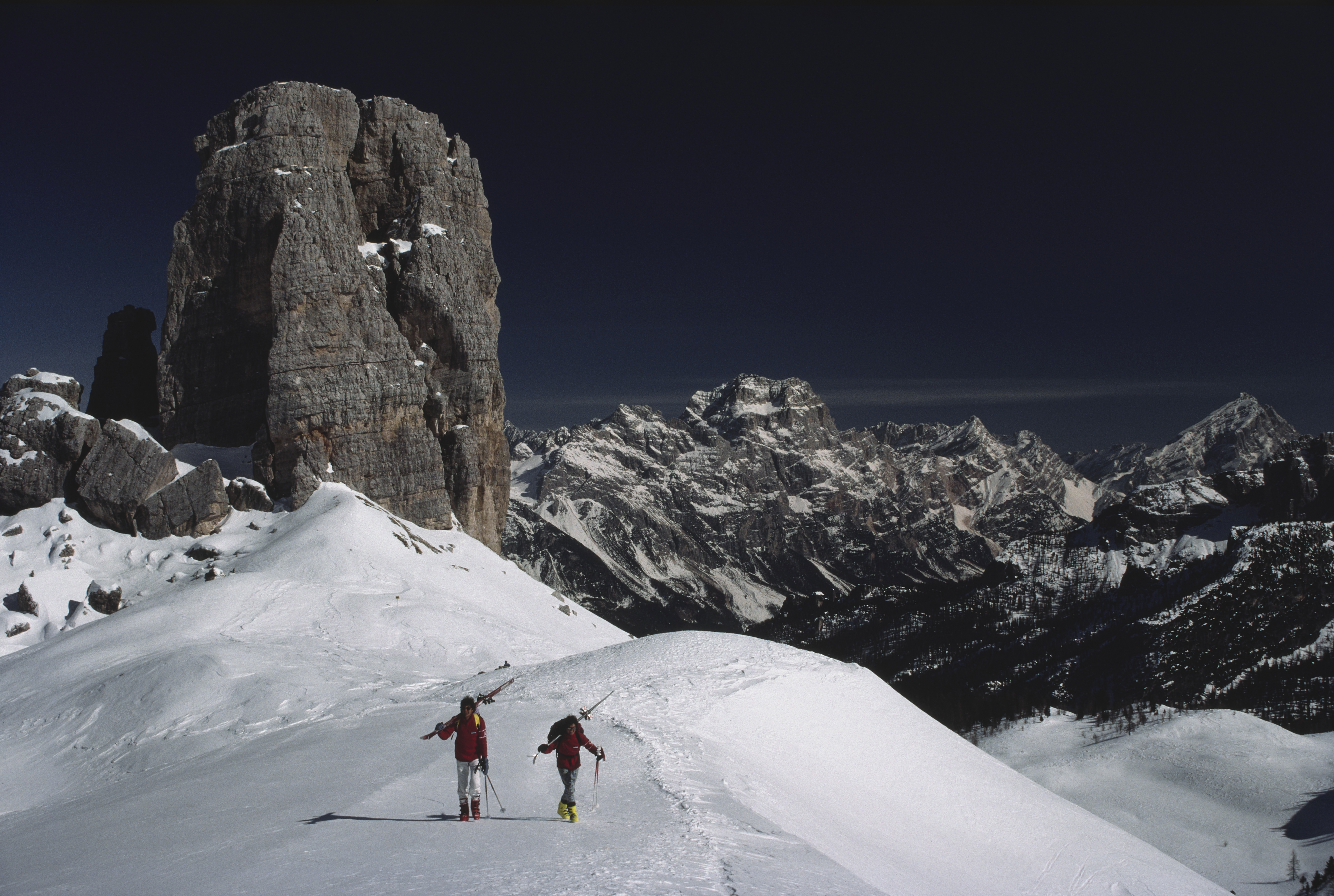 Two skiers walking along a narrow snow ridge in the Dolomites, carrying skis on their shoulders beneath towering limestone rock formations and a dark, dramatic winter sky.