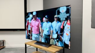 65-inch LG C4 TV photographed at an angle on a wooden stand. On the screen is an image of a golfer in a pink polo shirt.