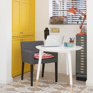 bright yellow cupboards behind home office workstation where laptop sits on a white table in front of a window with blinds