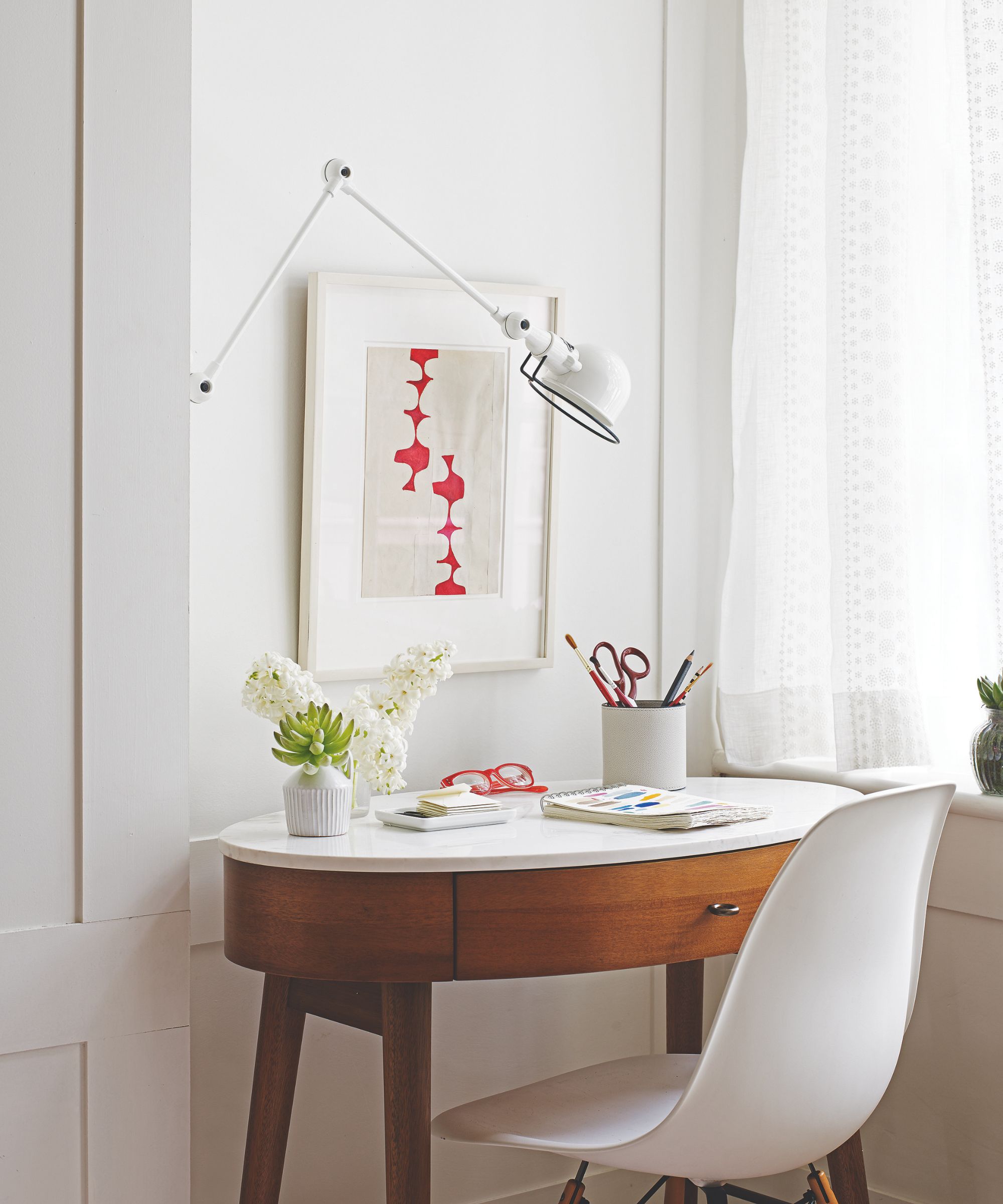 A white home office desk space with white walls, white sheer curtains, an oval-shaped wooden desk with white countertops, and white desk chair.