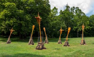 To Middle Species, With Love, Joyce Hwang, 2021. A series of wooden poles with wooden structures at the top of them, each held up by a pile of stones on a green grass.