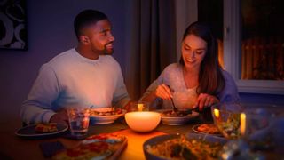 A couple eating dinner at a table in the dark with a Philips Hue Go smart lamp emiting bright warm light amongst their food in front of them.