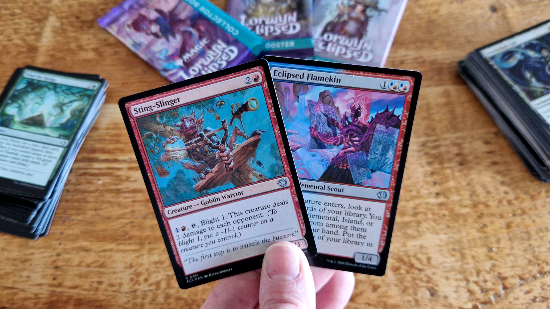 A hand holds up two cards in front of a wooden surface with decks and boosters laid out on it