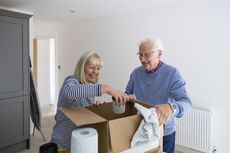 A senior couple unpacking a cardboard box filled with their kitchen belongings in the house they have just bought together. The woman is holding a mug while her husband is smiling and watching her.