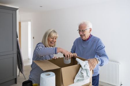 A senior couple unpacking a cardboard box filled with their kitchen belongings in the house they have just bought together. The woman is holding a mug while her husband is smiling and watching her.