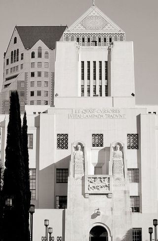 Black and white image of The Central Library, Los Angeles, 1926, stone status above the entrance, windows, tall conifer tree to the left, street lamps in a row either side of the drive way, pointed roof with Muriel design