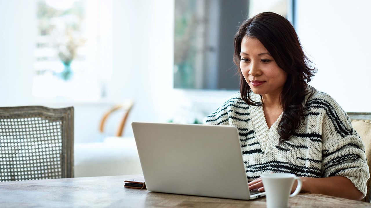 A woman sits at a dining room table with a laptop and a cup of coffee.