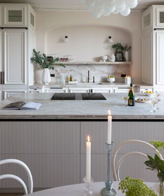 Neutral kitchen with pale pink painted walls, marble countertops, textured island, decorated with vases, plants, candles, glassware, large glass pendant with individual opal glass globes