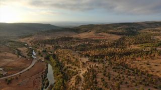 An aerial view of Morocco&#039;s hilly and reddish landscape that is spotted with trees by a river.