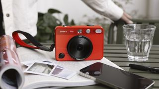 Leica Sofort 2 in red on a table resting on a book with instant prints scattered in foreground