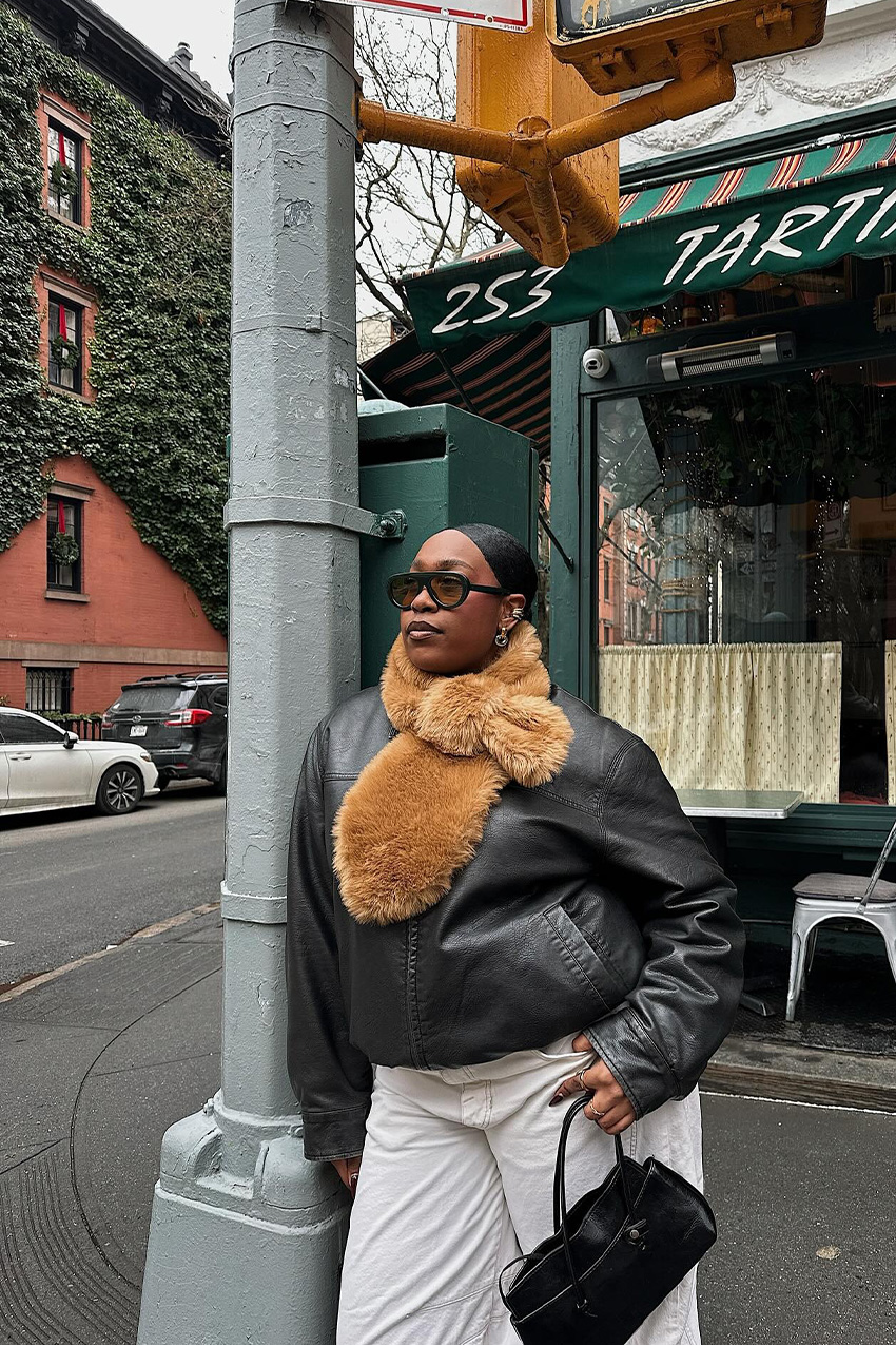 West Village winter trends are shown in a close street style photo of a woman leaning against a light pole in front of a restaurant in her winter outfit, wearing oversized two-tone aviator sunglasses with a black leather bomber jacket, white wide-leg jeans, a black shoulder bag, and a brown fur scarf