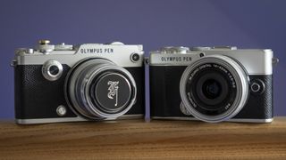 The Olympus PEN-F sat next to an Olympus PEN E-P7, on a wooden surface against a blue background
