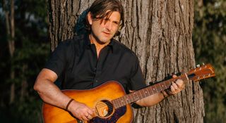 Christian Parker is photographed during golden hour, playing a Gibson acoustic against a tree.