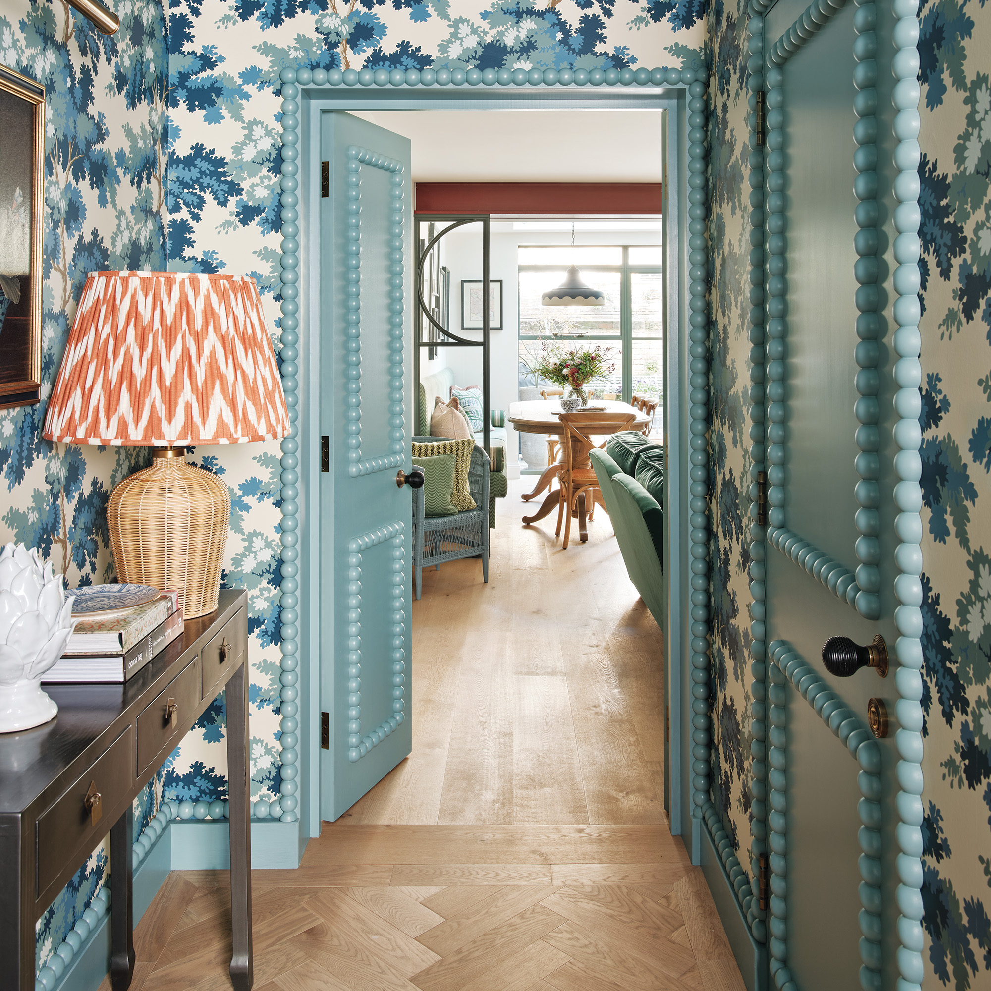 hallway with white walls with blue leaf-style print wallpaper blue doors with bobbin edges and panels and a wooden sideboard with rattan lamp and orange and white shade