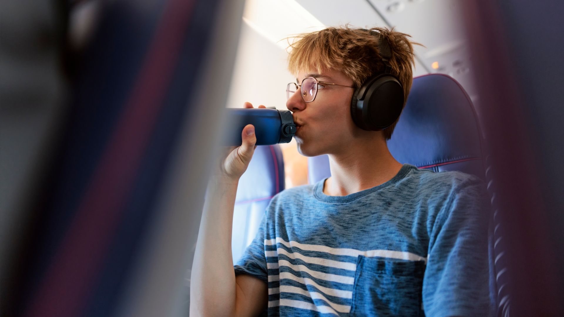 A boy with glasses drinking from a water bottle sitting on a plane