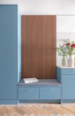 A bedroom with blue painted cupboards and bespoke seat with storage, a wood clad wall and mirror and a parquet pattern wood floor.