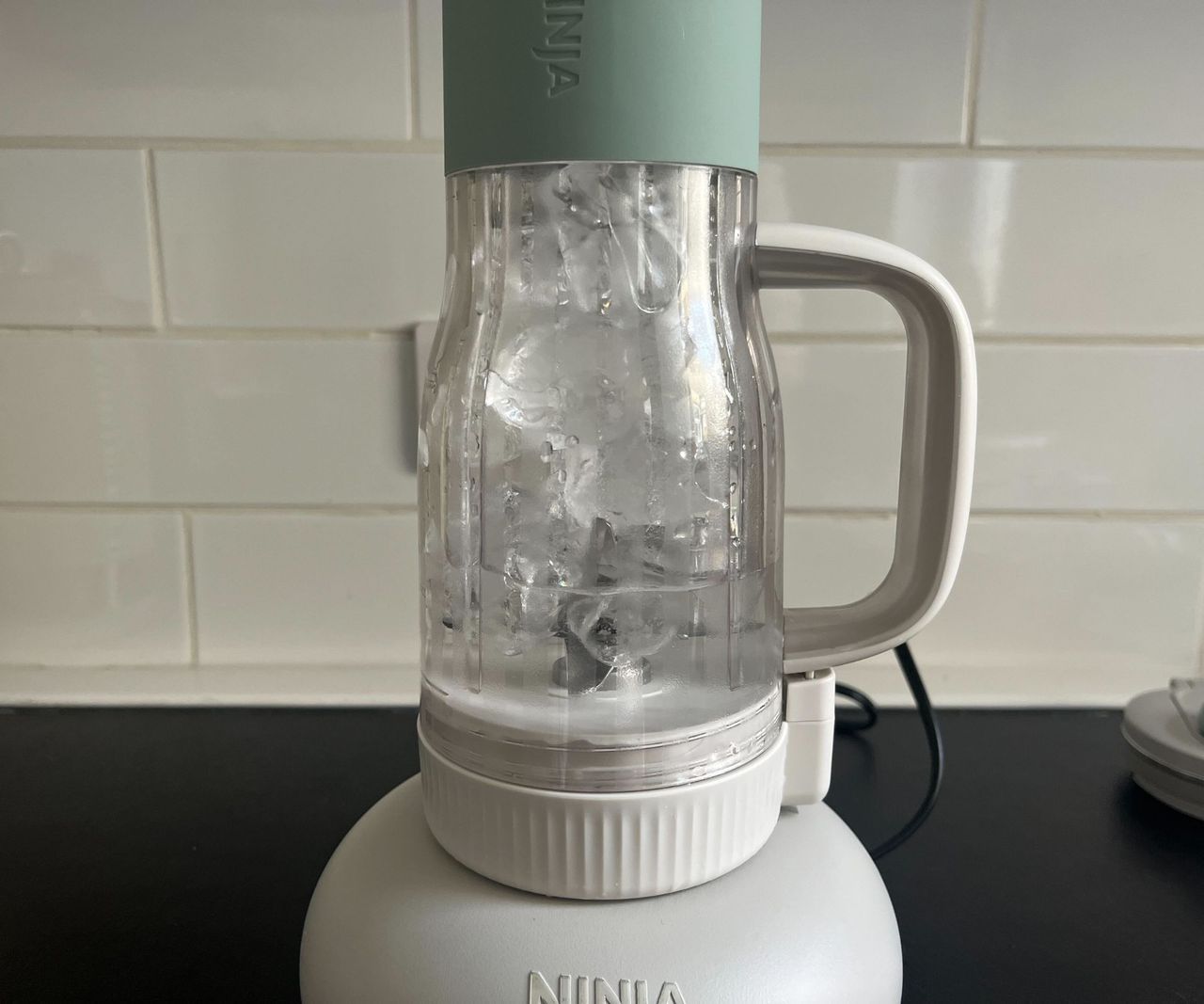 A blender tumbler filled with ice water, set against a white subway tile backsplash.