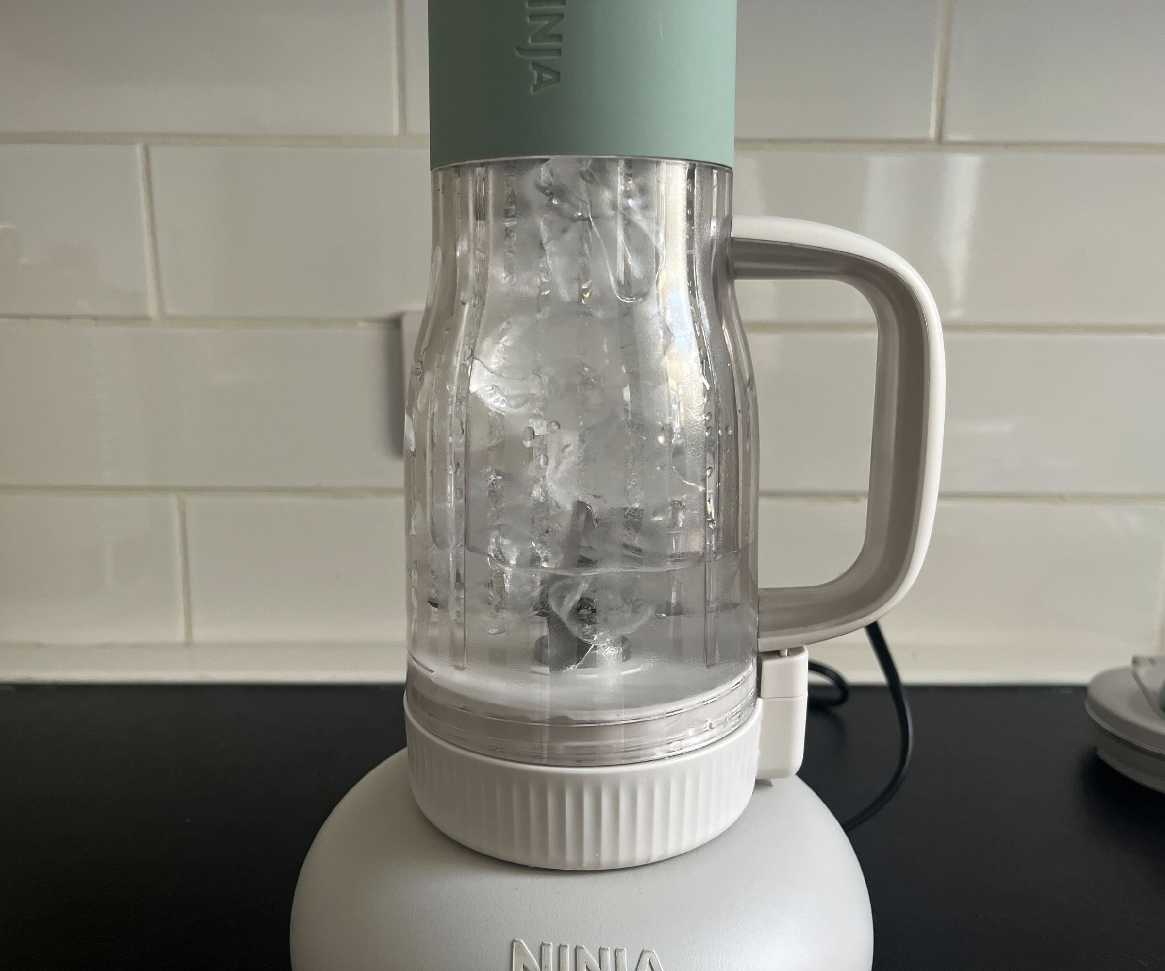 A blender tumbler filled with ice water, set against a white subway tile backsplash. 
