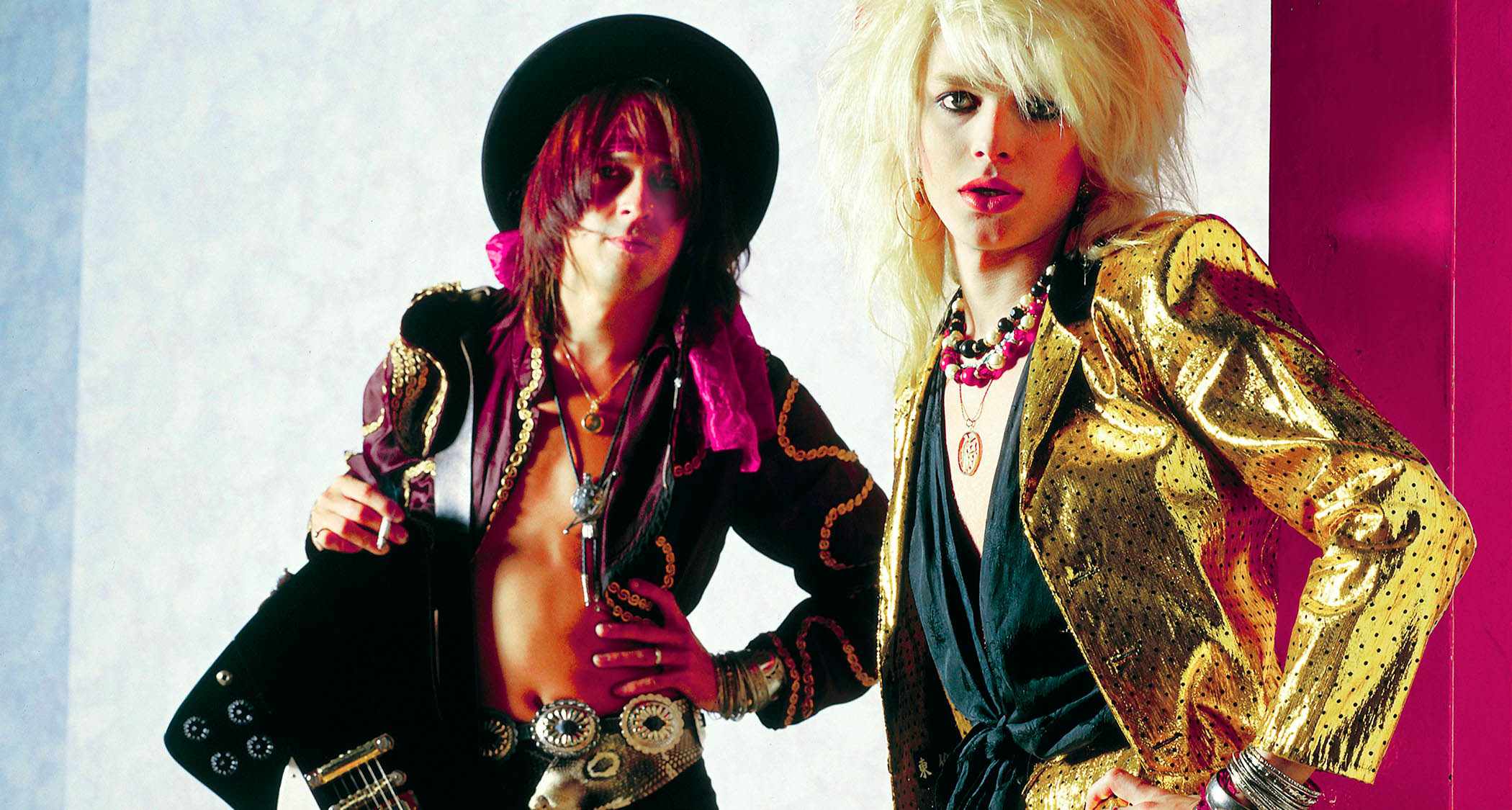Hanoi Rocks&#039; Andy McCoy [left] and Michael Monroe. Monroe wears a bright gold jacket and has blonde, &#039;80s rock-sized hair. McCoy is cradling a black Gibson Firebird and weats a wide-brimmed hat.