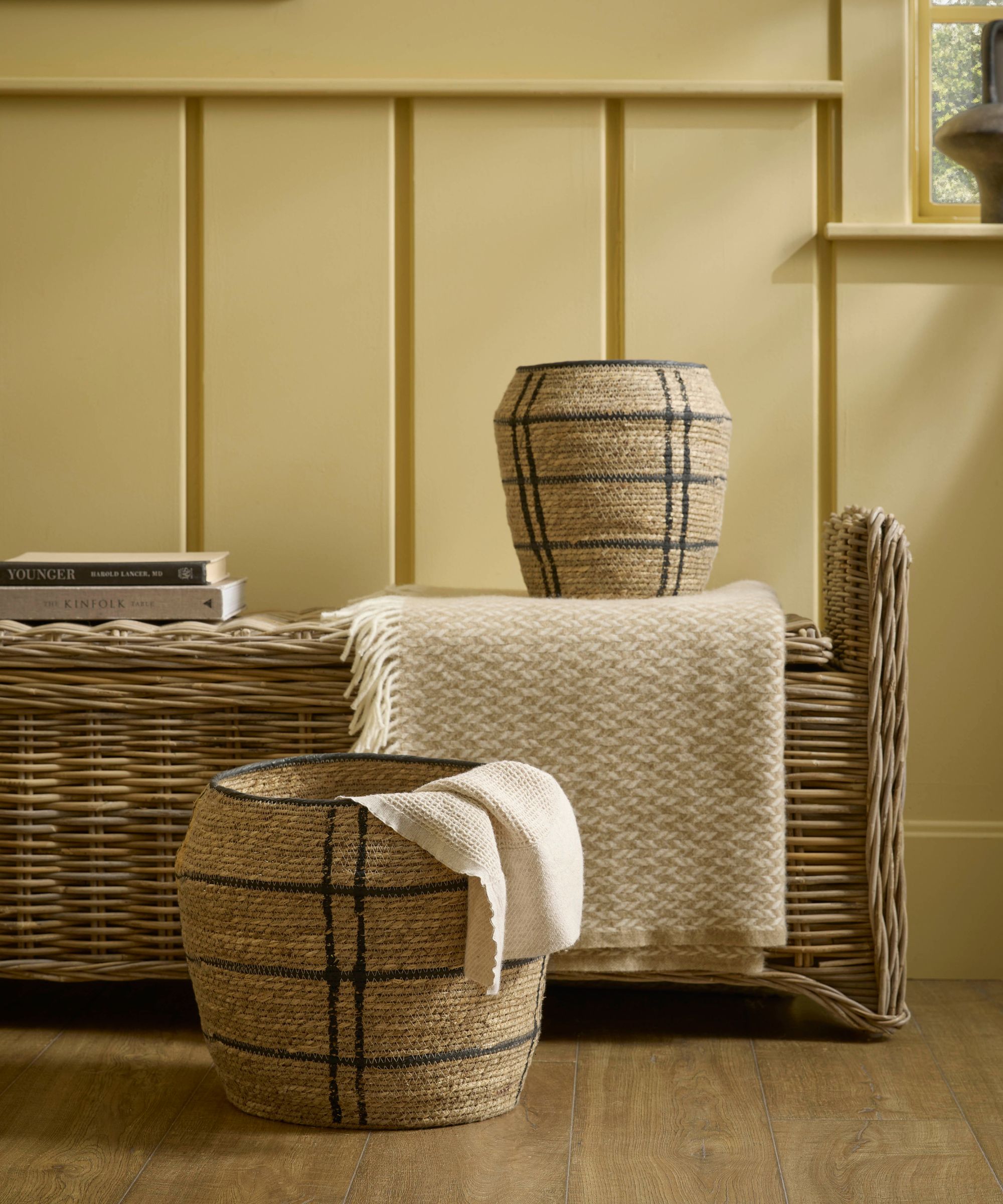 Two seagrass baskets with dark grid stripe detailing sit in front of a large rattan storage trunk.