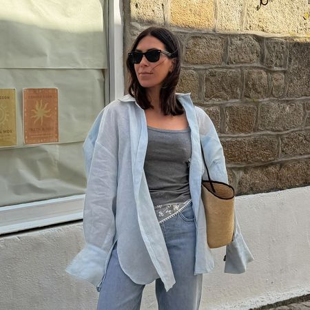 a woman wearing a blue oversized button-down shirt over a gray tnak top, blue jeans, a white lace scarf on her waist, a straw woven bag, and boat shoes