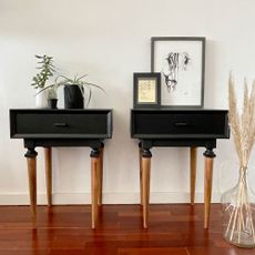 Two black vintage bedside tables on shiny wooden floor with with plant and 1 framed picture and 1 framed document on it