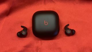 The Beats Fit Pro earbuds next to their charging case on a red backdrop