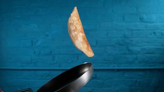 Pancake flipping in air from frying pan with blue brick wall behind.