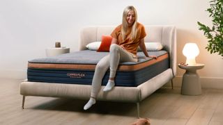 A blonde woman sitting at base of Brooklyn Bedding CopperFlex mattress on suede bed base.