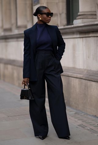 Hanukkah outfit idea is shown in a photo of a woman wearing a navy blazer with a navy turtleneck with matching trousers, gold earrings, a black bag, and black pointed heels