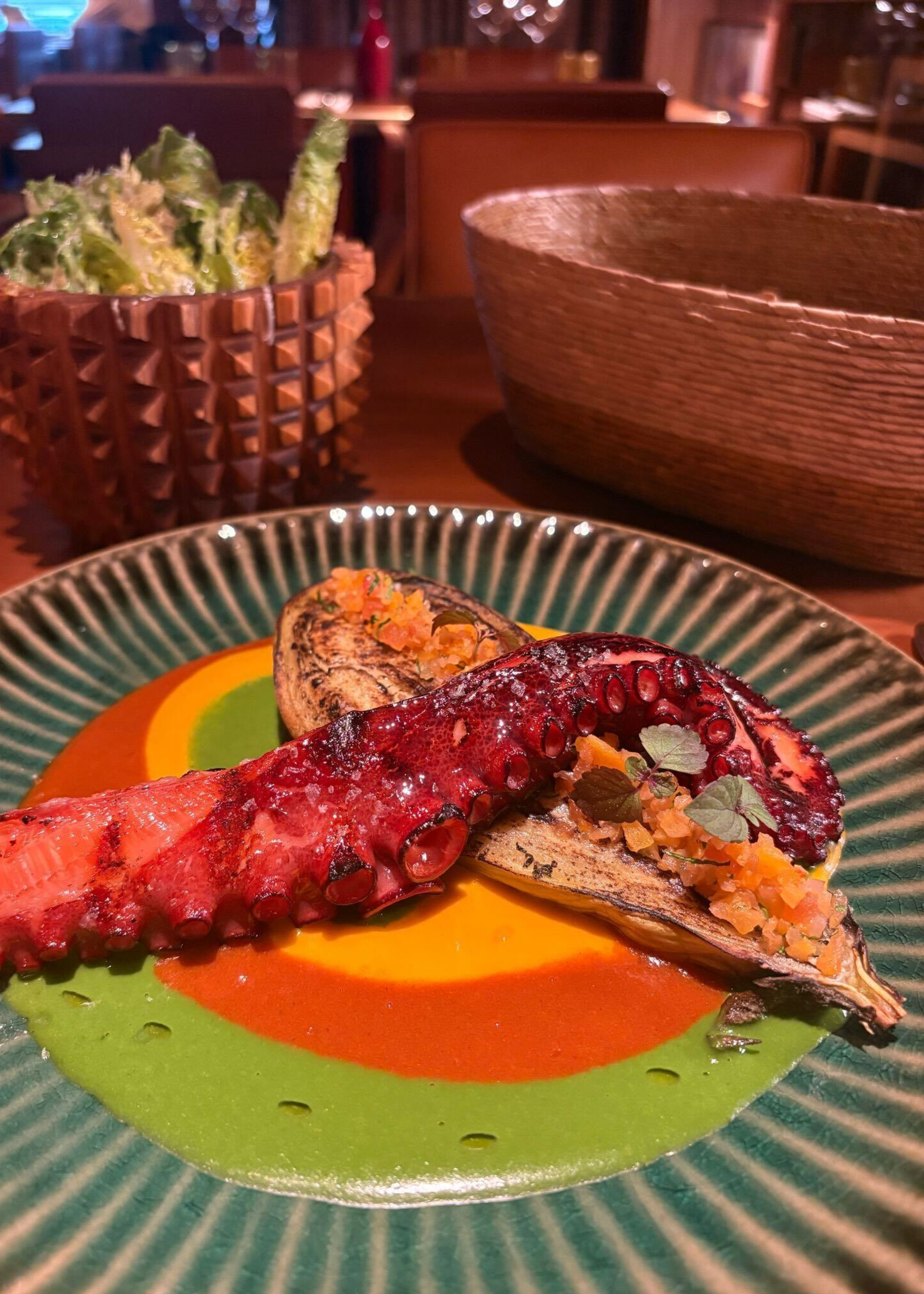 A plate of seared octopus and eggplant with a sobrassado sauce beside a bowl of Caesar salad, and a bread basket