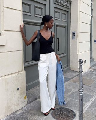 Scandinavian stylist and influencer Sylvie Mus poses in front of ornate doors on a Paris sidewalk wearing a low bun, black tank top, black tote bag, white linen pants, a denim jacket in her hand, and black sandals