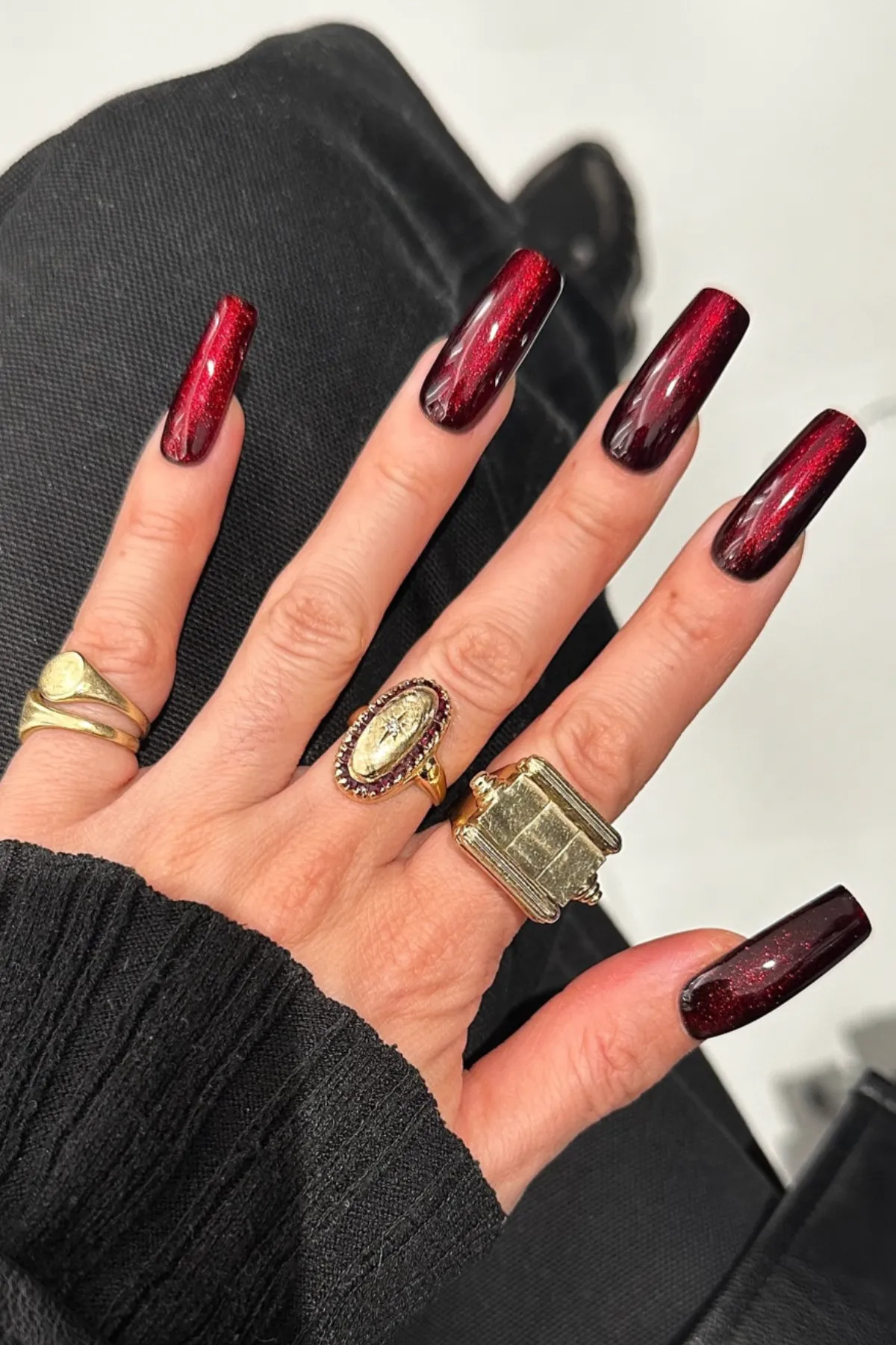 a close up of a woman's hand with gold rings wearing a metallic ruby red manicure