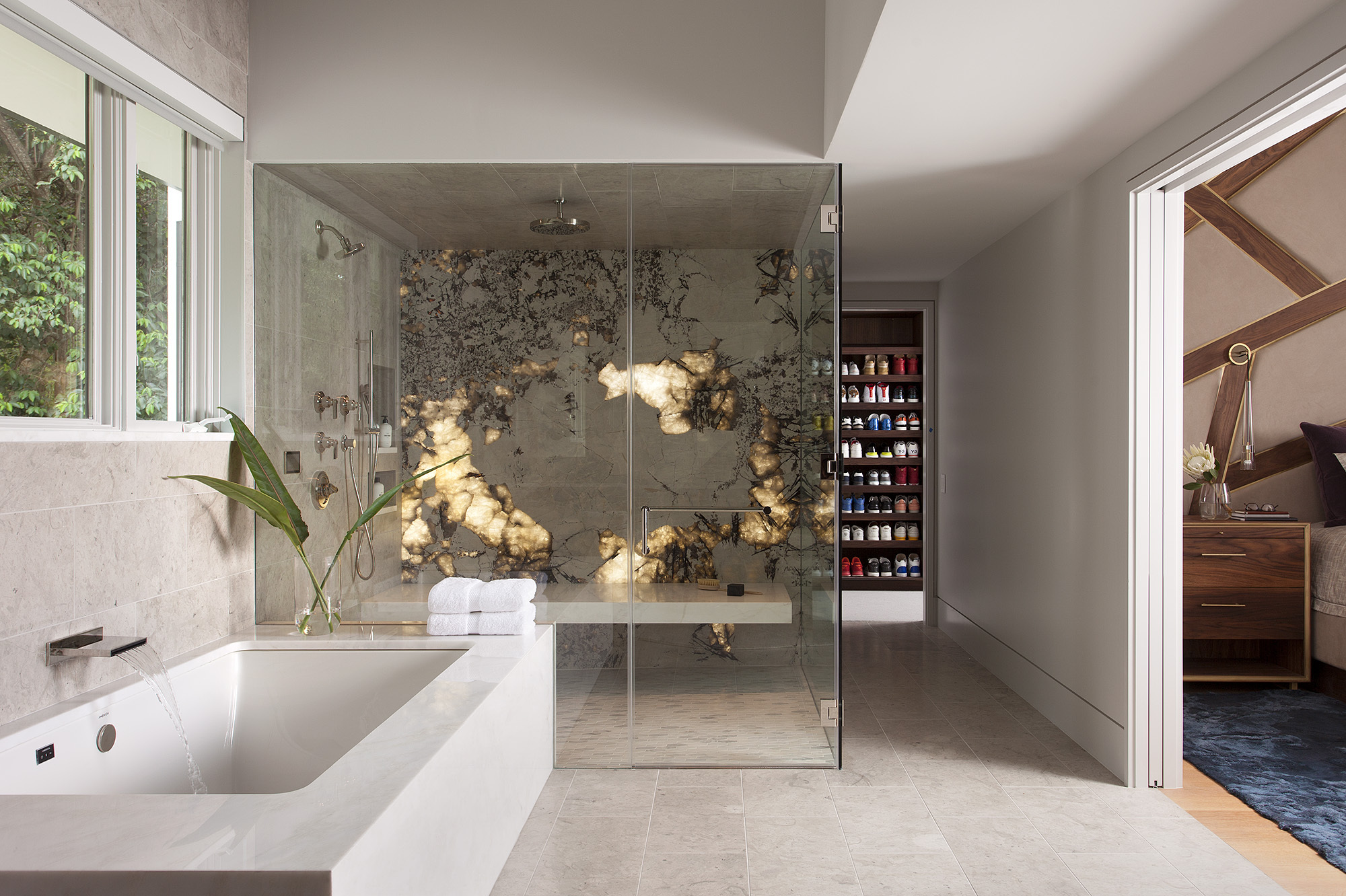 enclosed shower with backlit onyx wall with shower bench seat. elsewhere in the open bathroom is a built-in bath, and a doorway into the bedroom