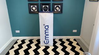 The Emma Original mattress shown in its box placed against a teal coloured wall and sat on a black and white rug