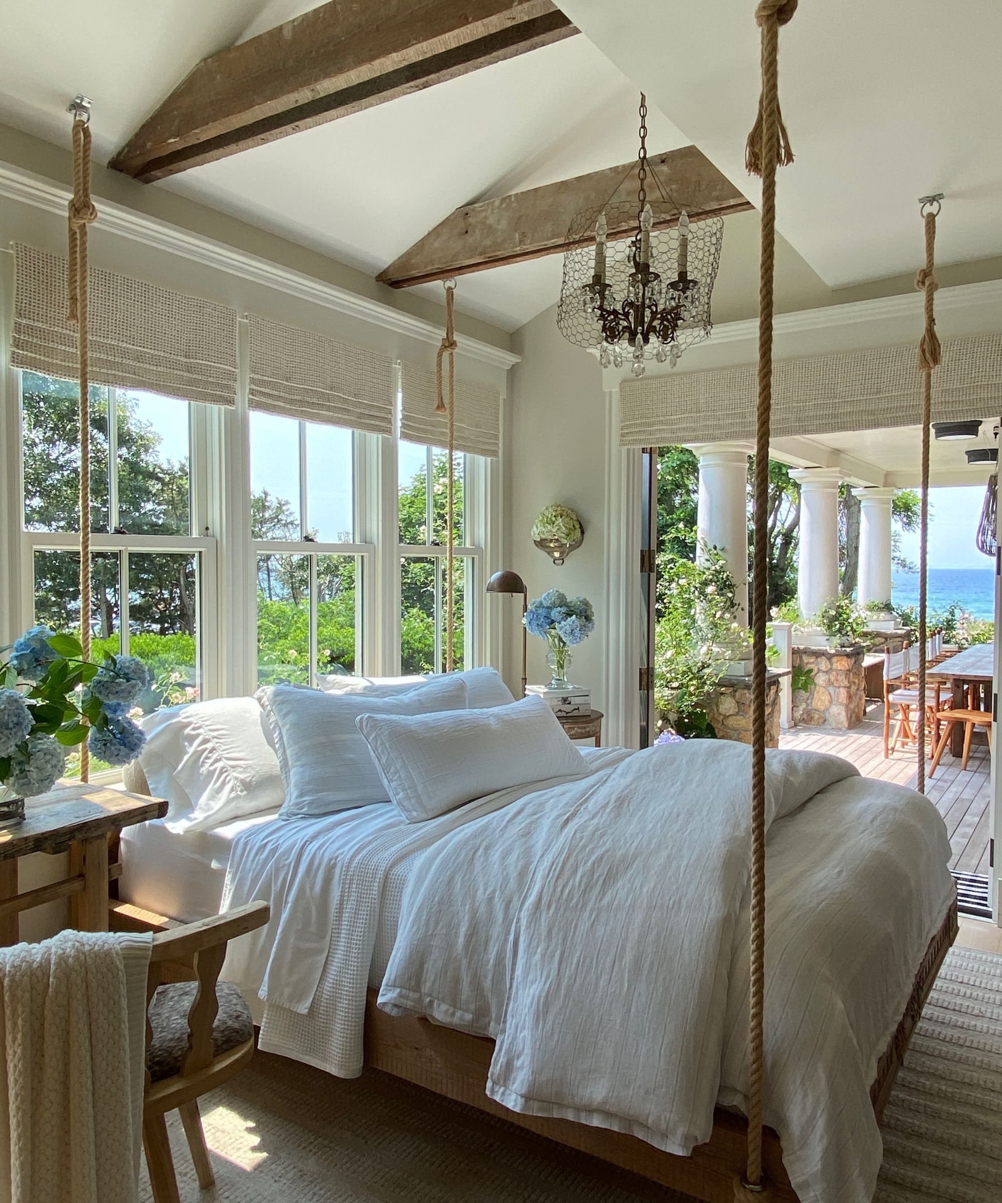 Sleeping porch with large swing bed with white linen bedding, wood flooring, chandelier onlooking porch with sea view