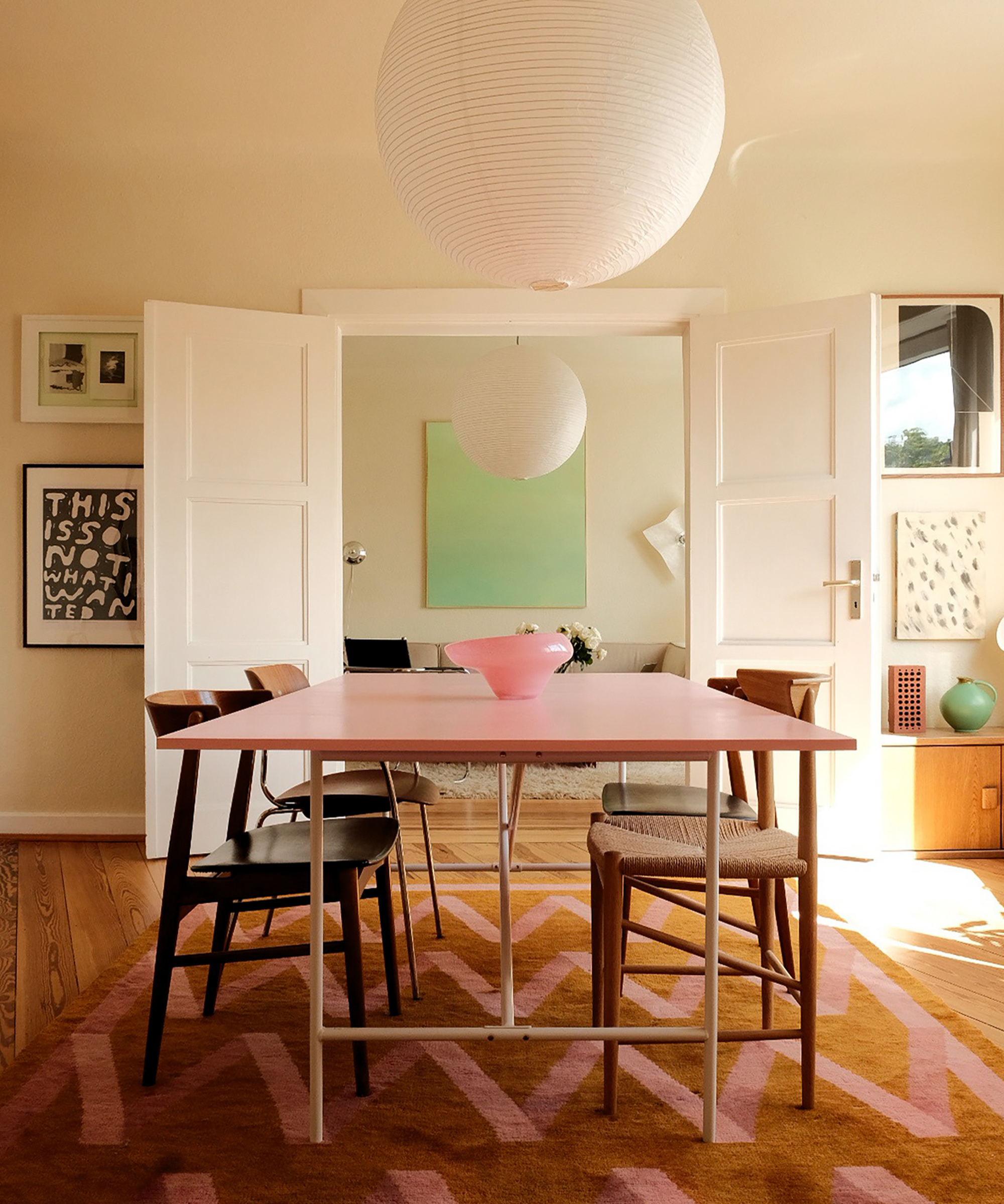 a dining room with a large pink dining table with chairs on either side, a paper pendant lamp, and a pink and brown zig zag rug on the floor