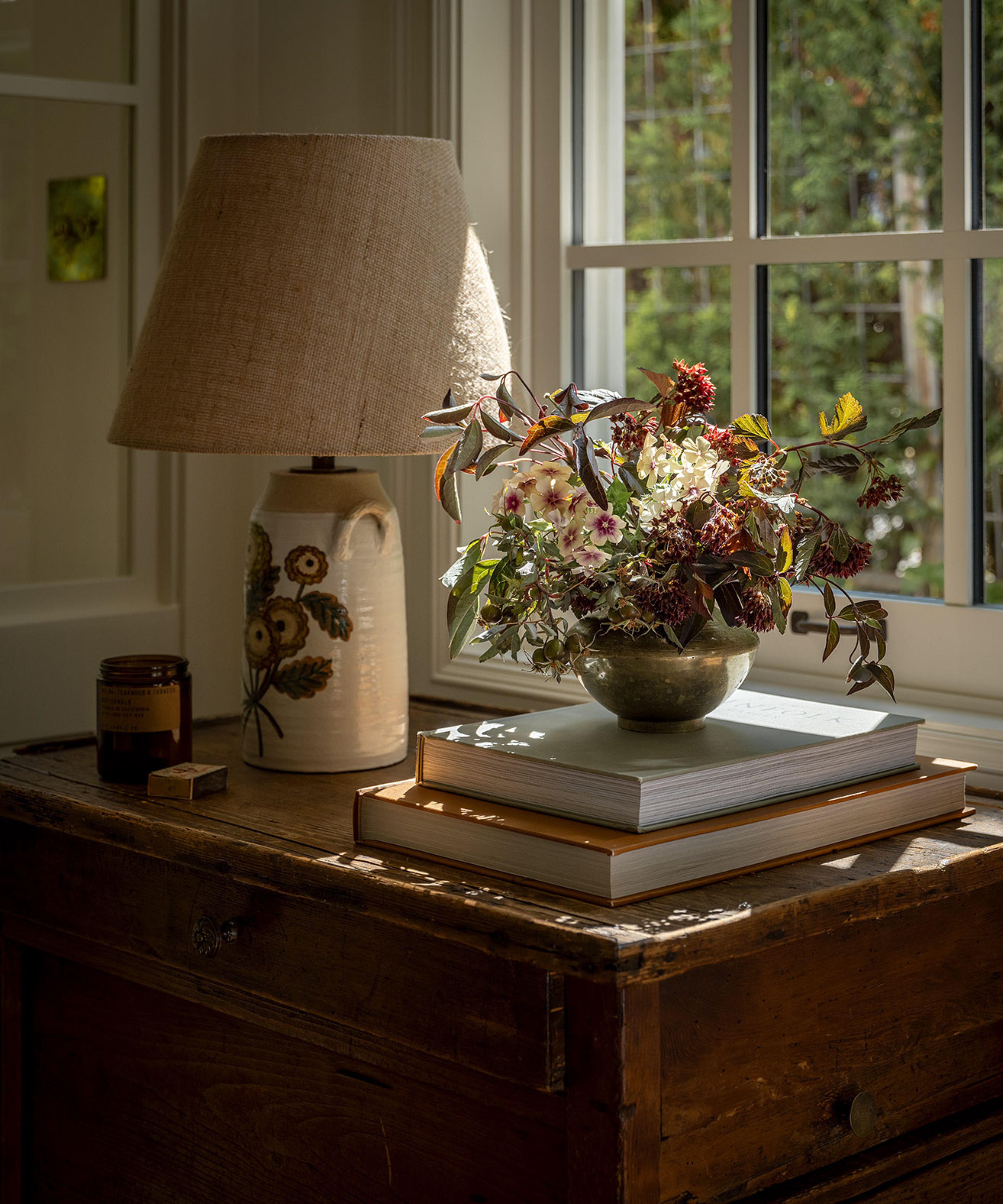 a detail shot in the Studio LeClercq garage DADU conversion of a rustic wooden unit in front of a window with a floral lamp base, and a pile of coffee table books with a vase of flowers